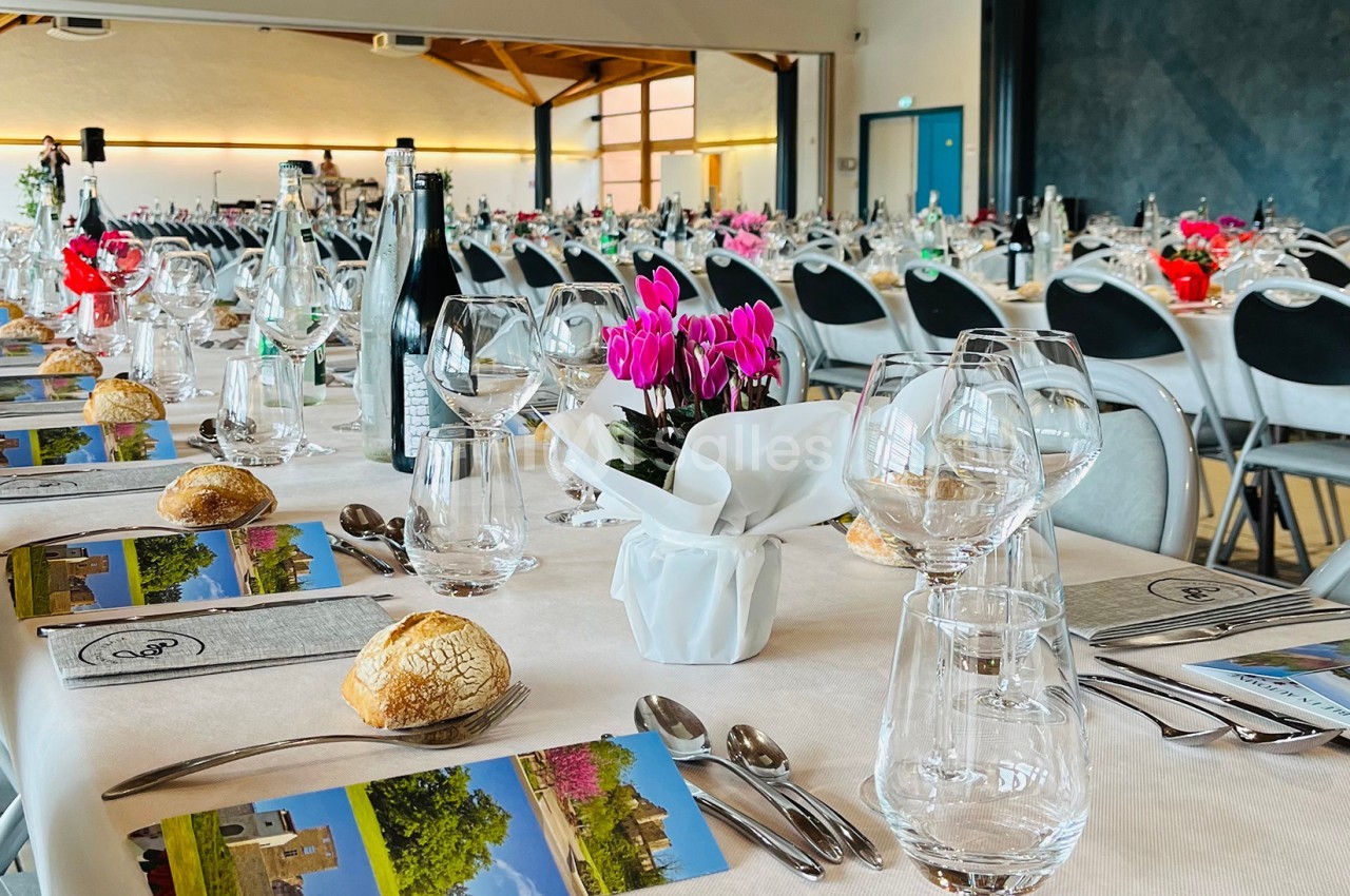 Tables dressées avec nappes blanches, couverts, verres, menus et décorations florales dans une grande salle lumineuse.
