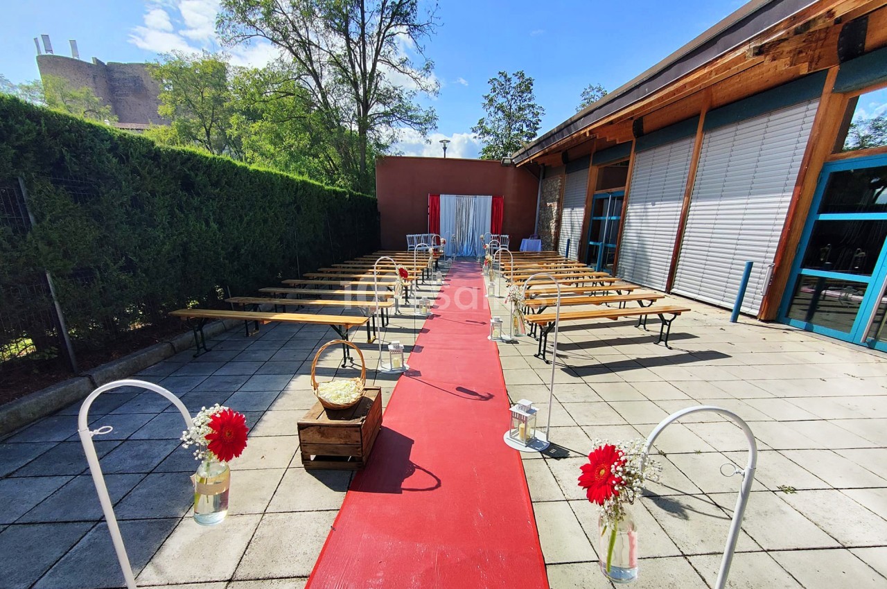 Allée centrale avec tapis rouge, bancs en bois de chaque côté, décorations florales et fond avec rideau blanc.