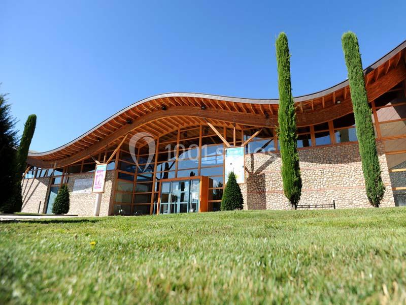 Façade d'un bâtiment moderne avec structure en bois, grandes baies vitrées et cyprès devant une pelouse verte.