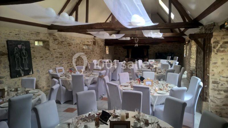 Salle de réception décorée pour un mariage, avec des tables rondes dressées et des chaises blanches sous des poutres…