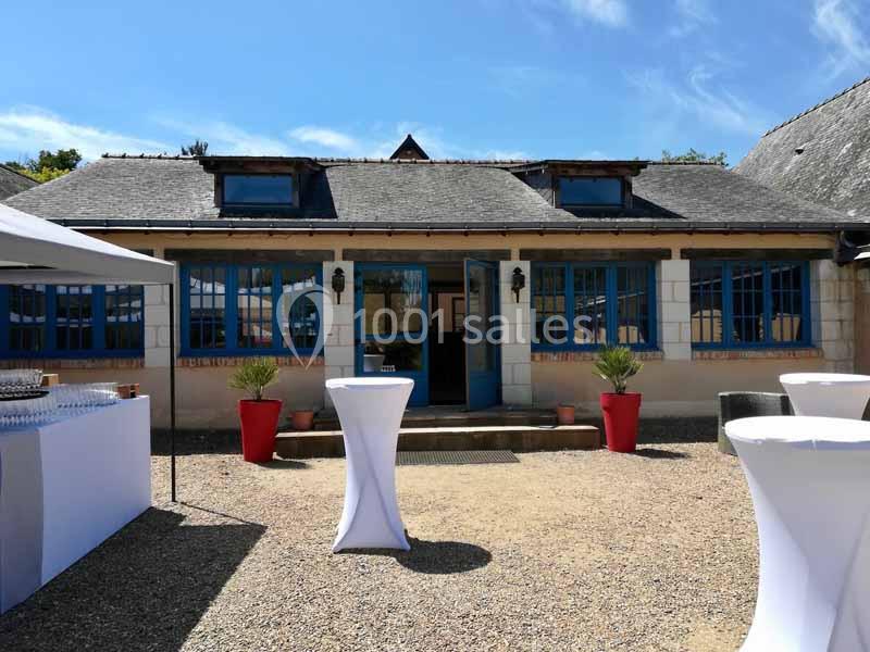 Façade d'un bâtiment avec fenêtres bleues, tables hautes blanches et parasols installés sur une cour en gravier.