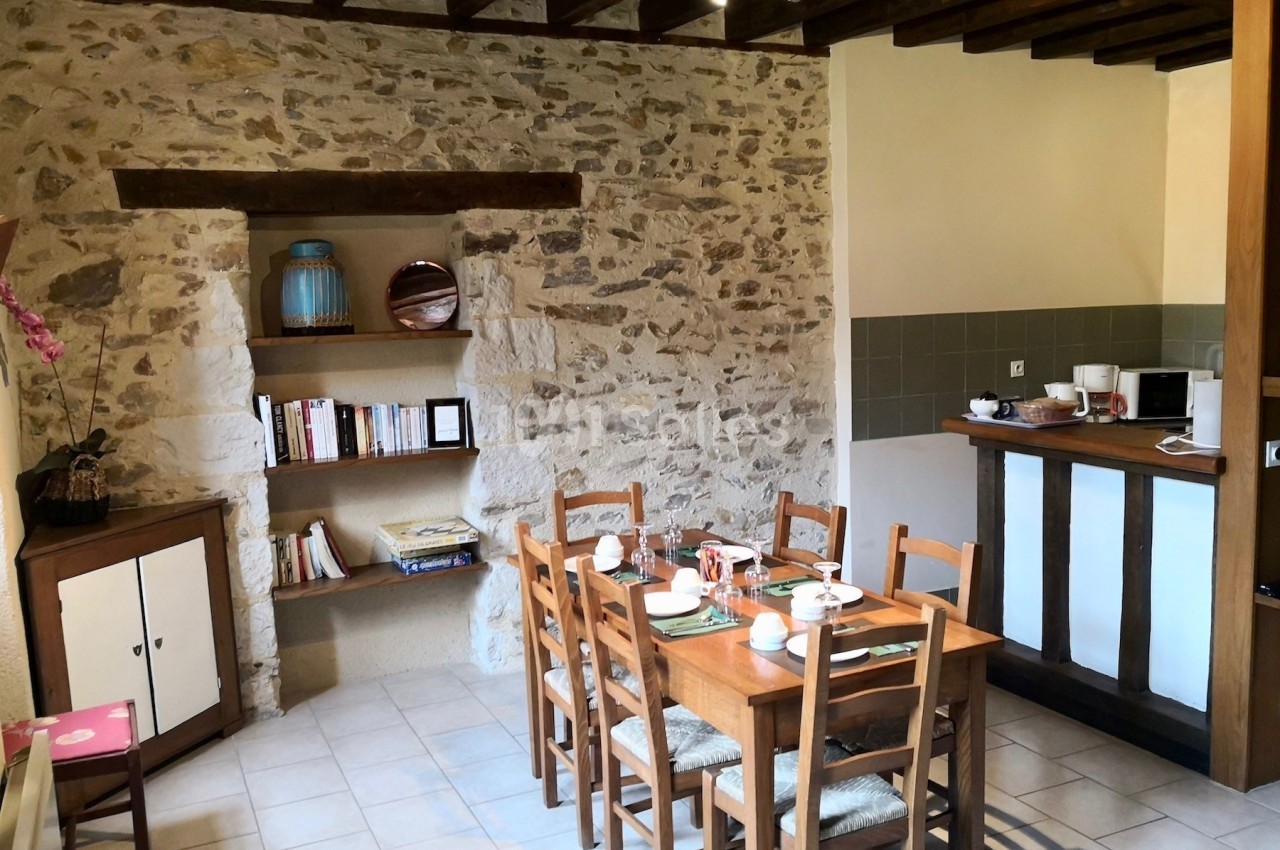 Salle à manger rustique avec table en bois dressée, mur en pierre apparente, étagères intégrées et cuisine ouverte.
