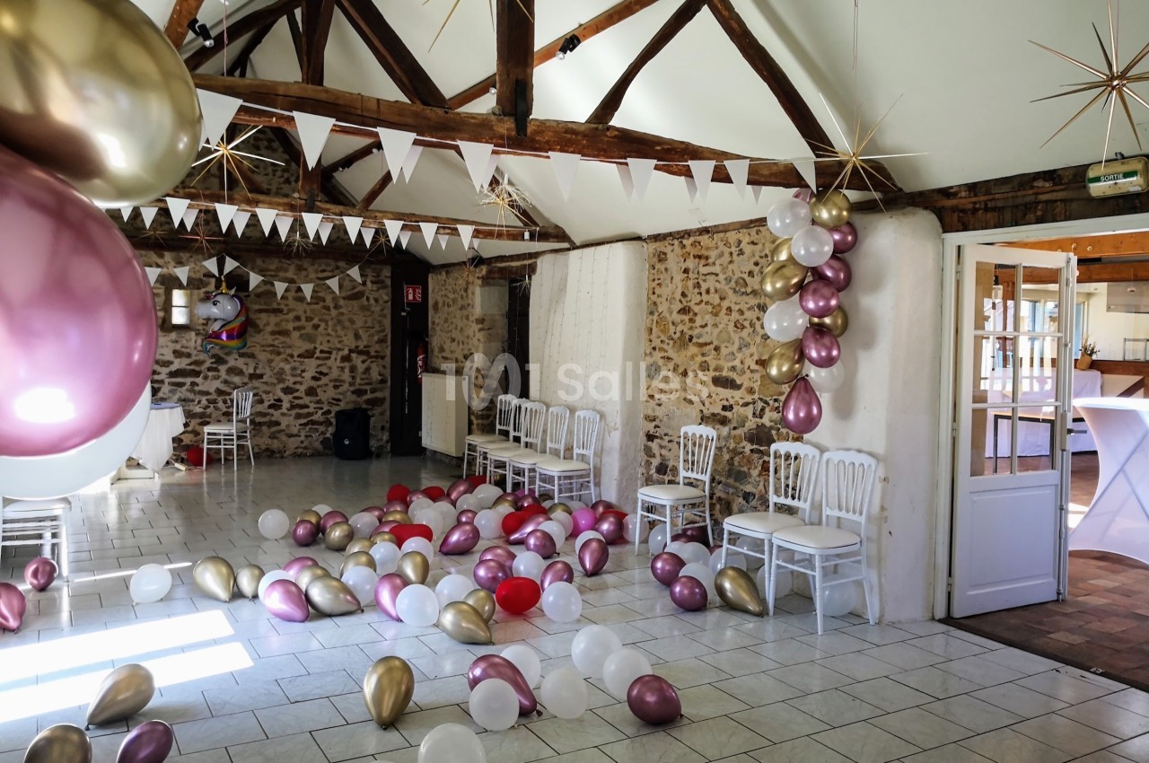 Salle décorée pour une fête avec des ballons colorés au sol, des guirlandes et des chaises alignées contre un mur en pierre.