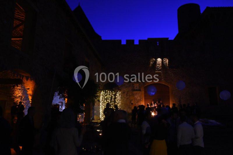 Cour d'un château éclairée par des guirlandes lumineuses et des lanternes, avec des invités rassemblés en soirée.