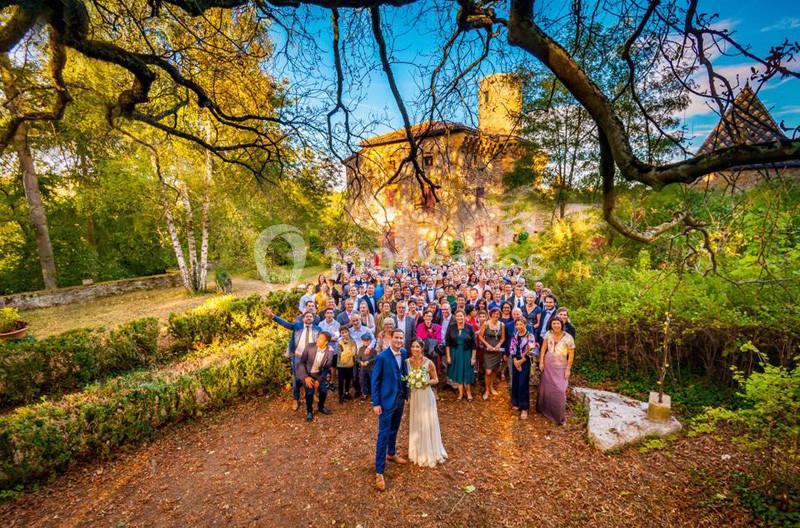 Groupe de personnes posant pour une photo de mariage en extérieur, devant un bâtiment ancien entouré de verdure.