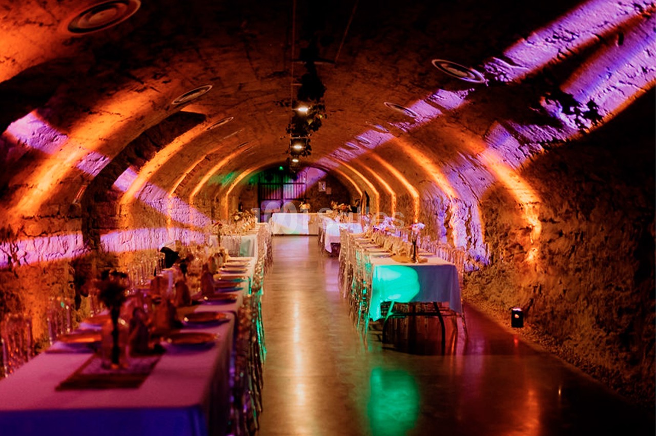 Salle voûtée en pierre éclairée par des lumières colorées, aménagée avec des tables dressées pour un événement.