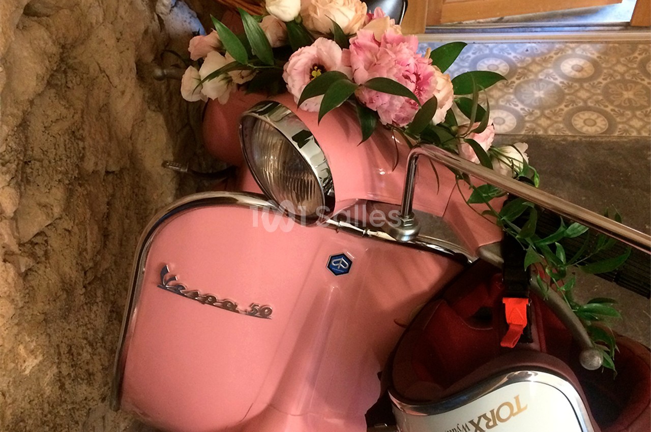 Scooter rose décoré de fleurs roses et blanches, stationné près d'un mur en pierre et d'une porte en bois.