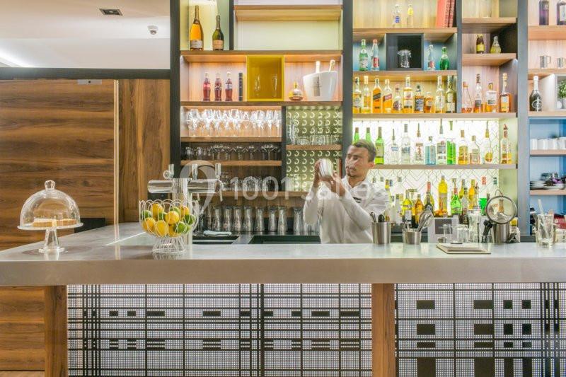 Un barman prépare un cocktail derrière un comptoir moderne, avec des étagères remplies de bouteilles et de verres.