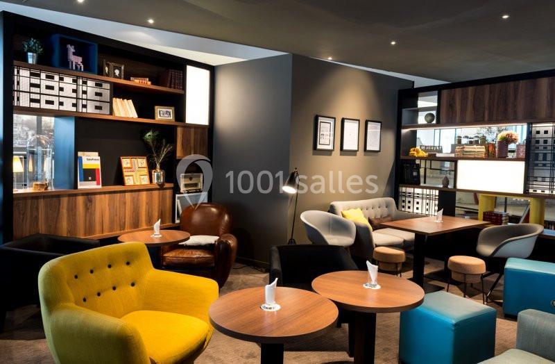 Espace lounge moderne avec fauteuils colorés, tables en bois, étagères décoratives et éclairage tamisé.