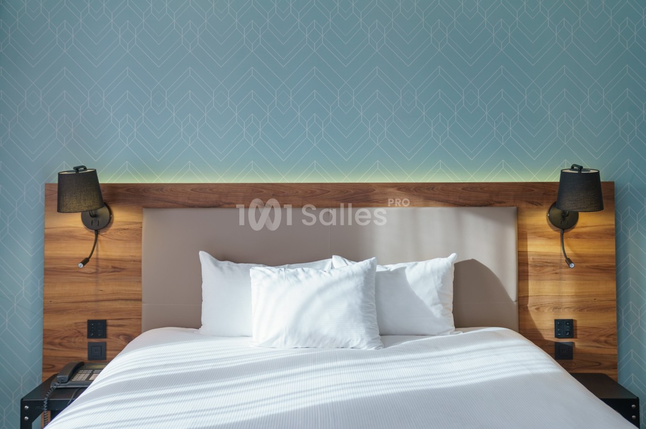 Lit double avec tête de lit en bois, deux lampes murales noires et un mur bleu à motifs géométriques en arrière-plan.
