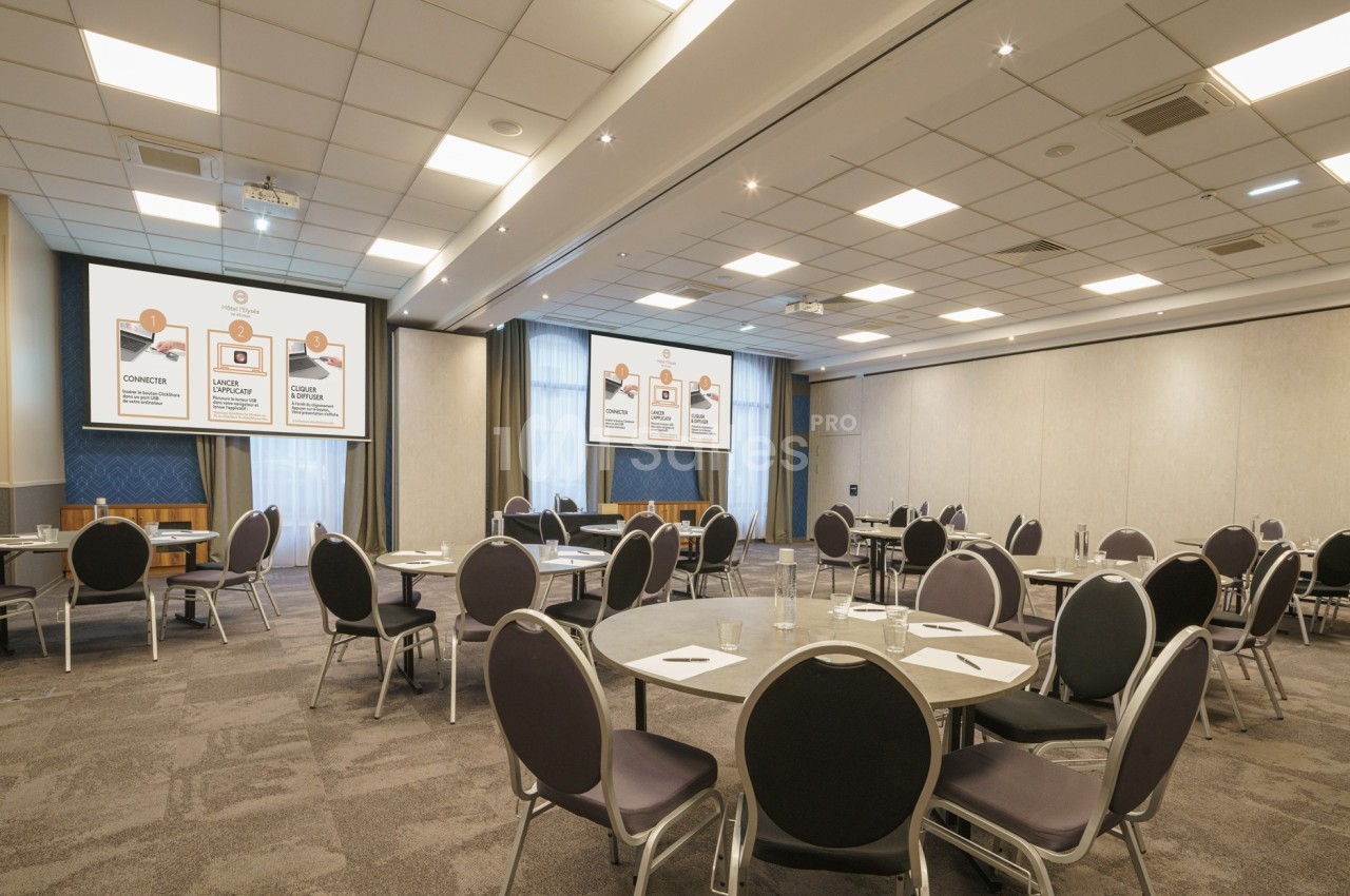 Salle de conférence lumineuse avec tables rondes, chaises noires et écrans de présentation affichant des diapositives.