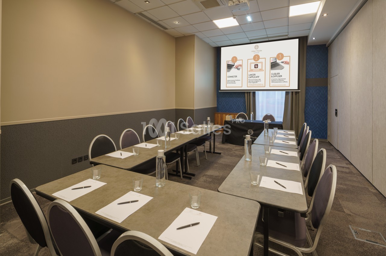 Salle de réunion équipée de tables en U, chaises, blocs-notes, bouteilles d'eau et écran de présentation allumé.