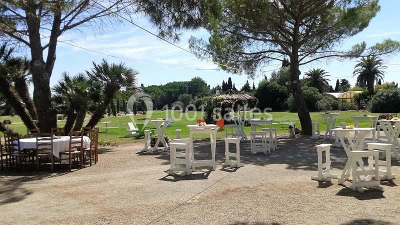 Espace extérieur avec tables et chaises blanches disposées sous des arbres, donnant sur une pelouse et des palmiers.