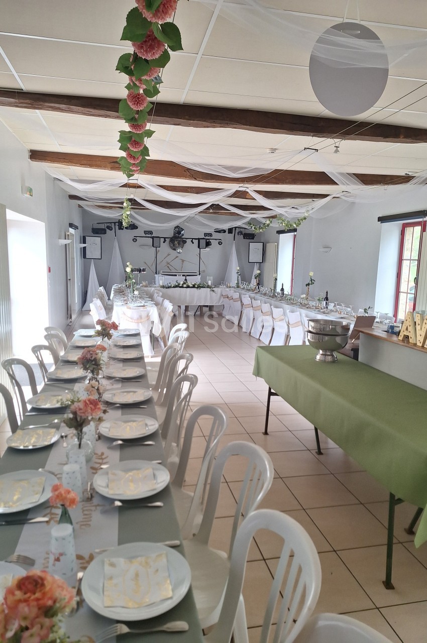 Salle décorée pour un événement avec tables dressées, nappes vertes, chaises blanches et guirlandes suspendues.