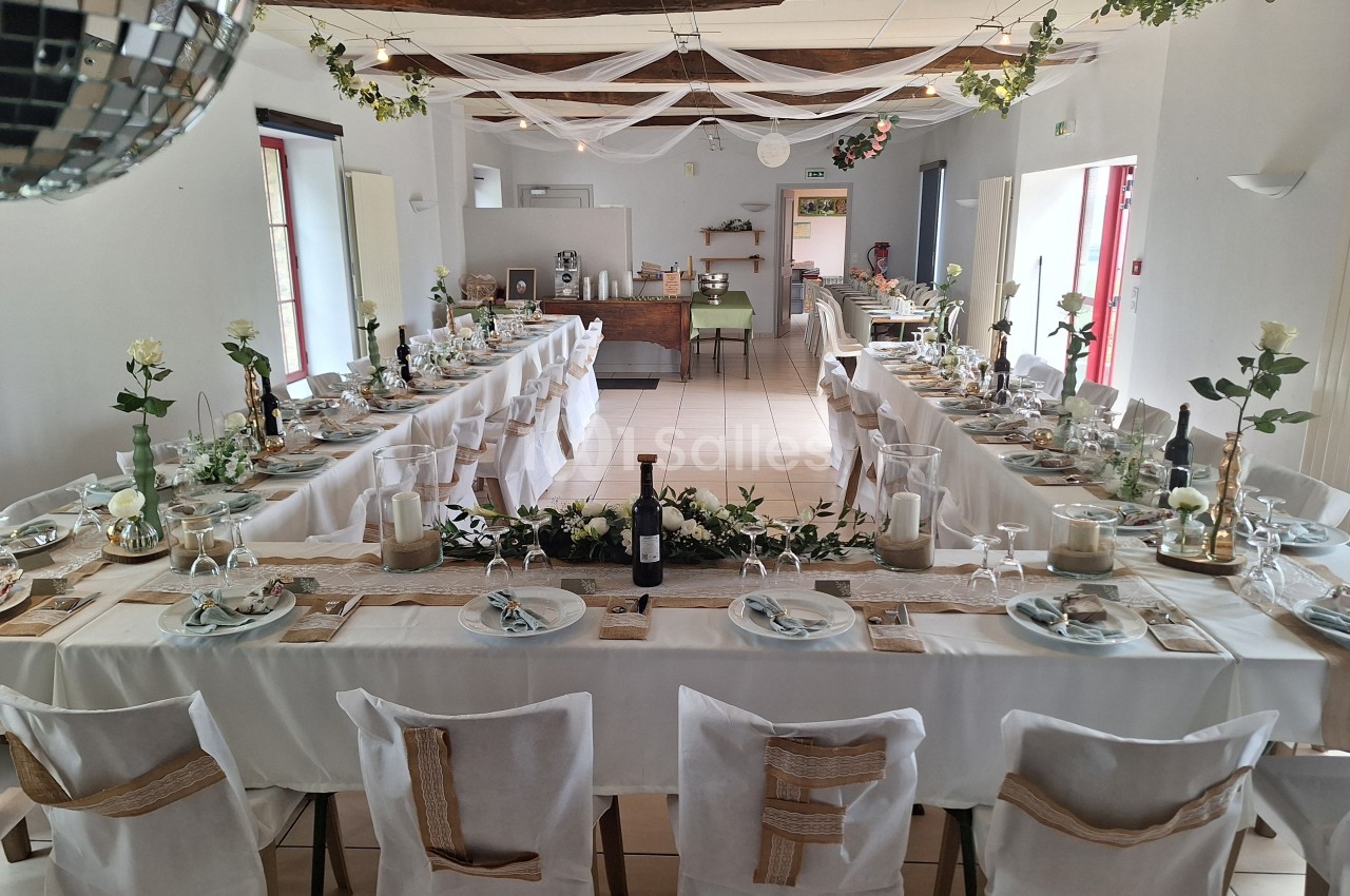 Salle décorée pour un repas festif avec longues tables dressées, nappes blanches, fleurs et guirlandes suspendues.