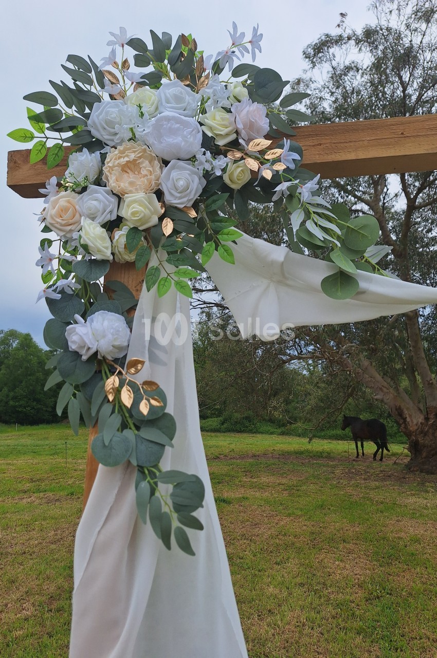 Arrangement floral blanc et vert avec des touches dorées, drapé de tissu blanc, fixé à une structure en bois dans un champ.