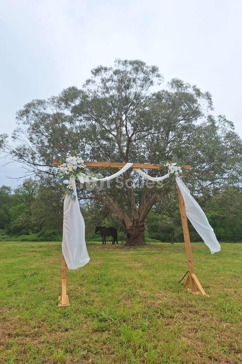 Arche de mariage en bois décorée de voilages blancs et de fleurs, installée dans un champ devant un grand arbre.
