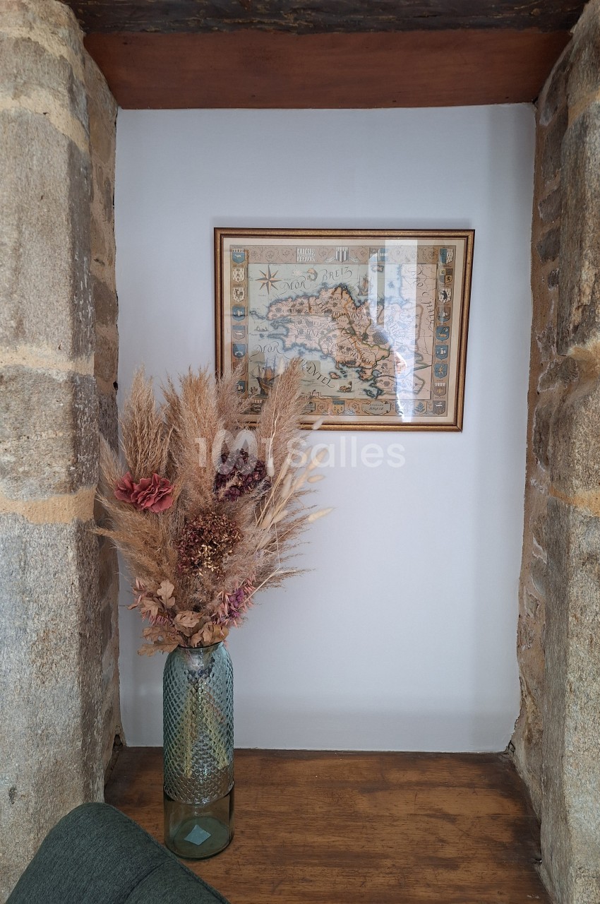 Vase en verre vert contenant un bouquet de fleurs séchées, posé sur une étagère entre deux murs en pierre, avec une carte…
