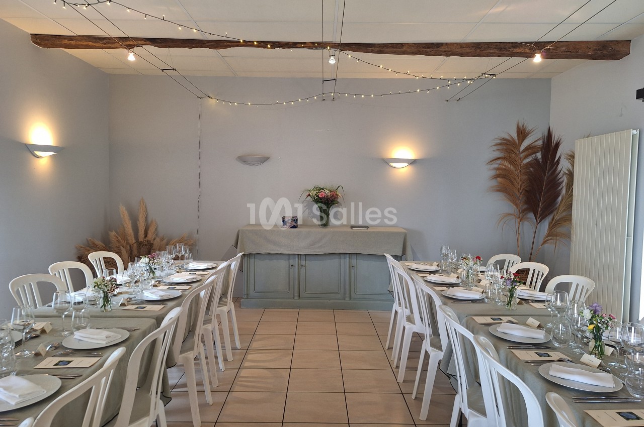 Salle décorée pour un repas, avec tables alignées, nappes beiges, chaises blanches et bouquet de fleurs sur un buffet.