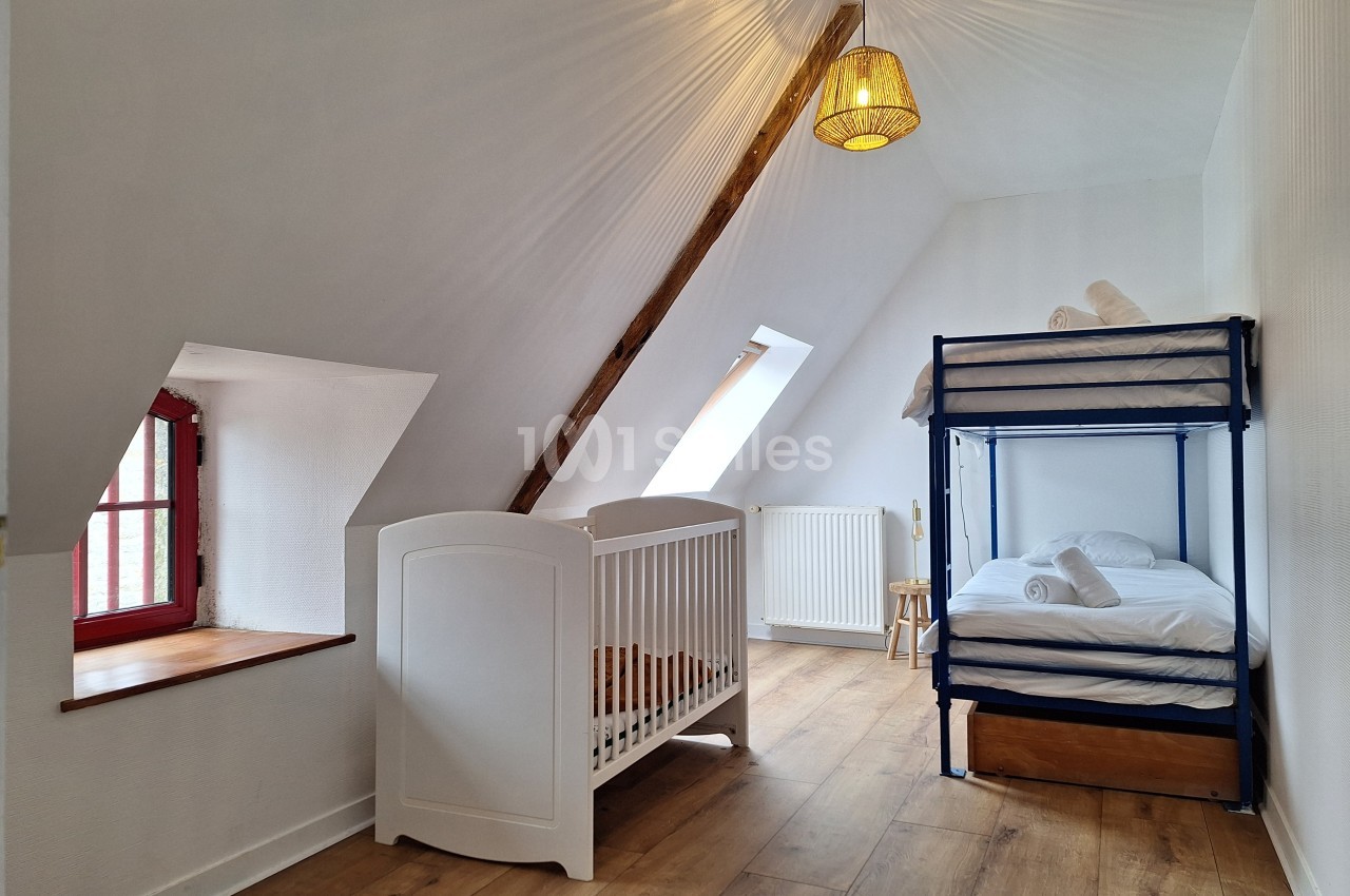 Chambre mansardée avec lit superposé, lit bébé, fenêtre à battants et parquet clair.