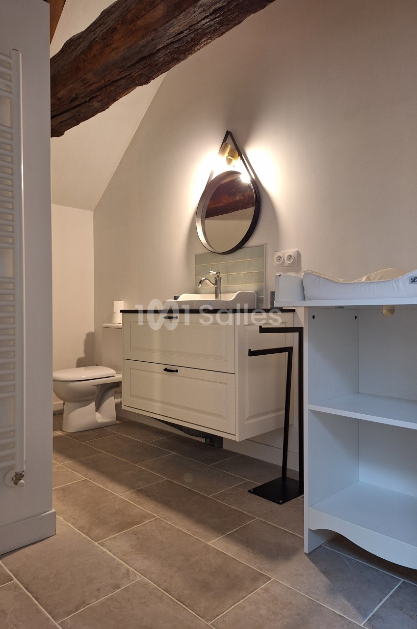 Salle de bain avec meuble blanc, lavabo, miroir mural triangulaire, table à langer et poutre apparente au plafond.