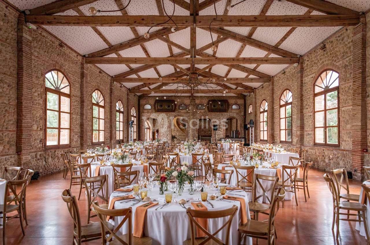 Salle de réception en briques avec poutres apparentes, tables rondes dressées et grandes fenêtres laissant entrer la lumière.
