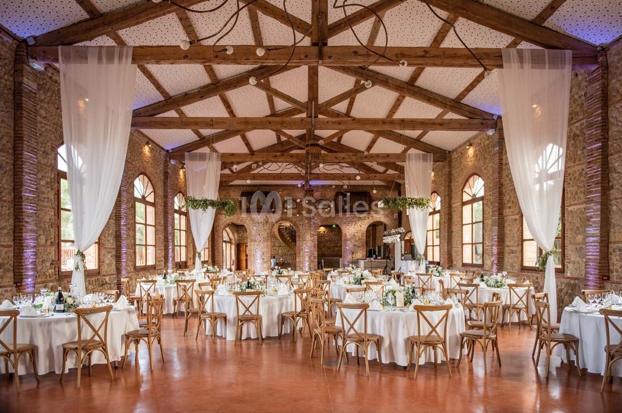 Salle de réception spacieuse avec tables rondes décorées, chaises en bois et grandes fenêtres en arc.