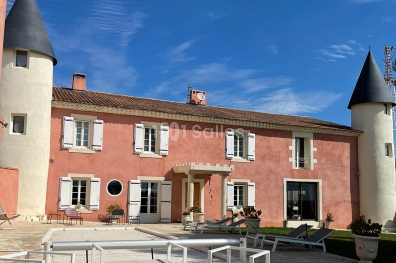 Façade d'une maison rose de style château avec deux tours, entourée d'une terrasse et de mobilier extérieur.