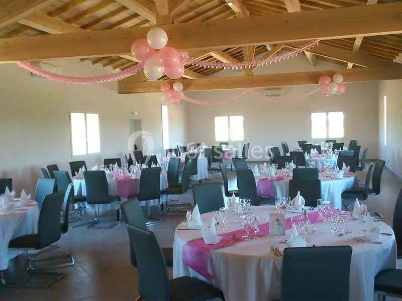 Salle de réception décorée avec des tables rondes, nappes blanches, chemins de table roses et ballons suspendus.