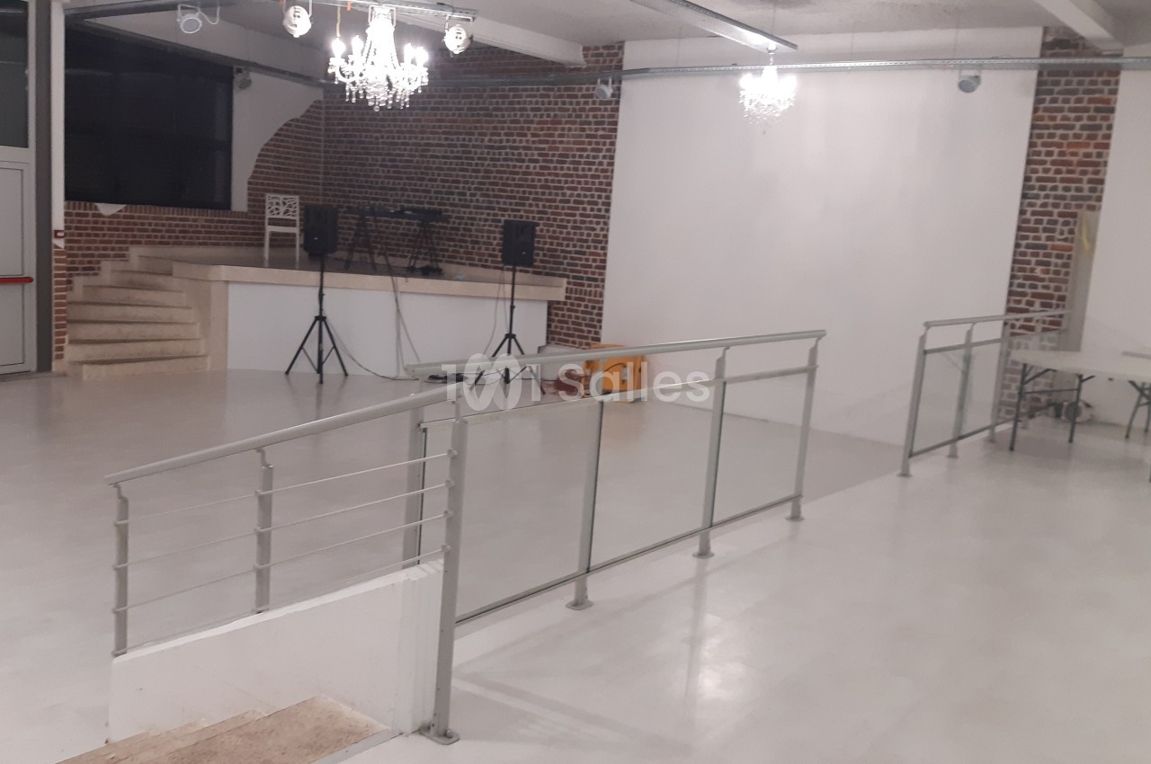 Salle vide avec sol clair, murs en briques, rampe d'accès, estrade avec enceintes et éclairage suspendu.