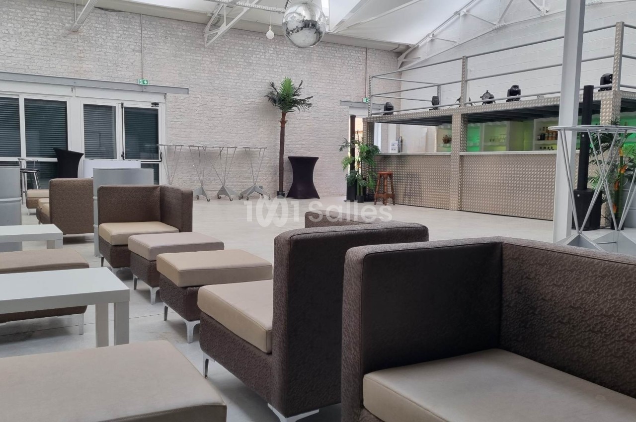 Salle lumineuse avec canapés, tables basses, bar moderne et décoration minimaliste, sous un plafond en verrière.