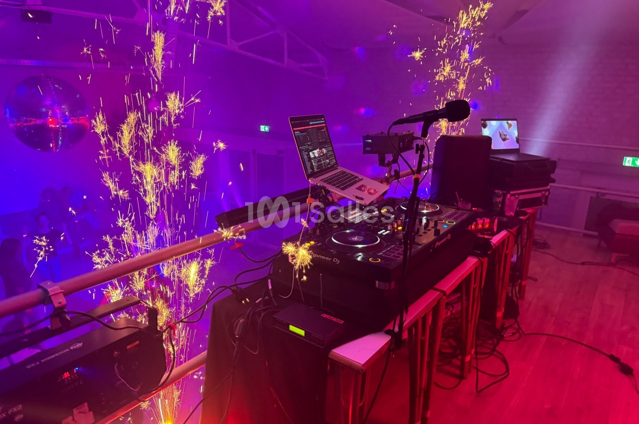 Poste de DJ avec platines et ordinateur, entouré d'effets pyrotechniques dans une salle éclairée par des lumières colorées.
