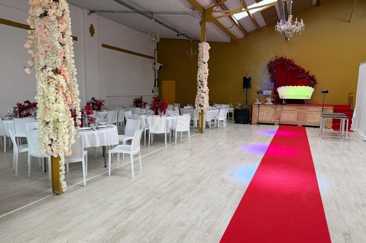 Salle de réception décorée avec des tables blanches, des colonnes fleuries et un tapis rouge menant à une estrade.