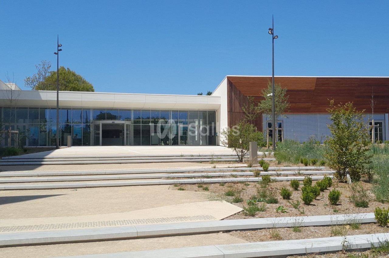 Entrée d'un bâtiment moderne avec façade vitrée et revêtement en bois, entourée de végétation et d'allées pavées.