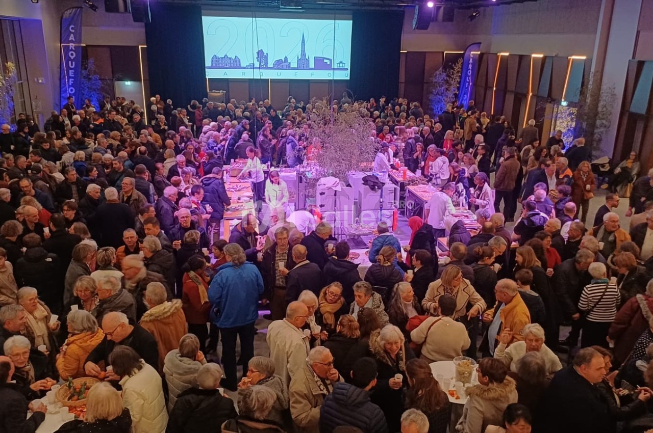 Foule rassemblée dans une grande salle éclairée, avec un buffet central et un écran projetant une image en arrière-plan.