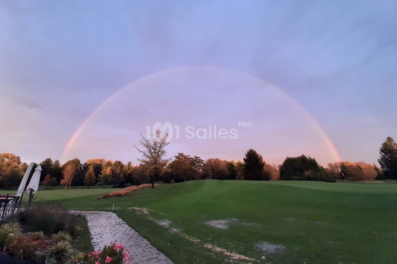 Un arc-en-ciel au-dessus d'un terrain de golf verdoyant bordé d'arbres sous un ciel légèrement nuageux.