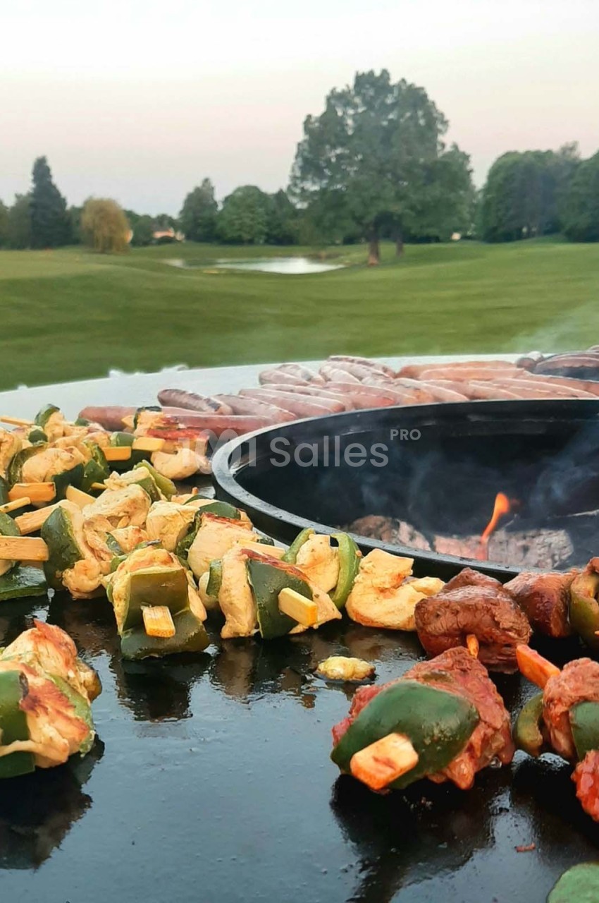 Brochettes de légumes et de viande grillant sur une plancha ronde, avec un parc verdoyant en arrière-plan.