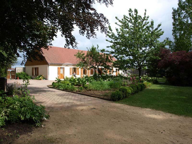 Maison à un étage avec toit en tuiles rouges, entourée d'un jardin arboré et d'une allée pavée.