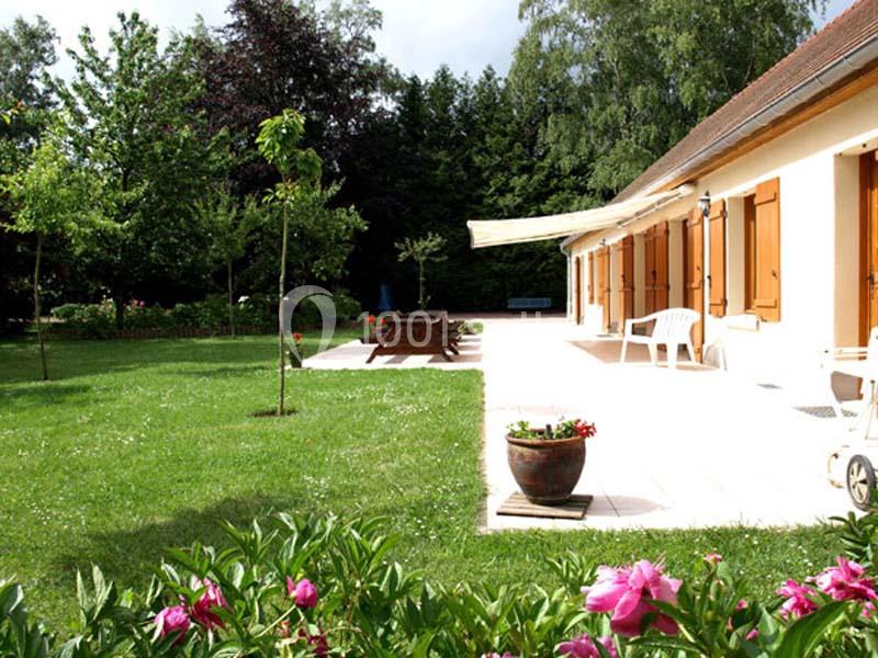 Jardin avec pelouse, fleurs, arbres, terrasse ensoleillée et maison aux volets en bois.