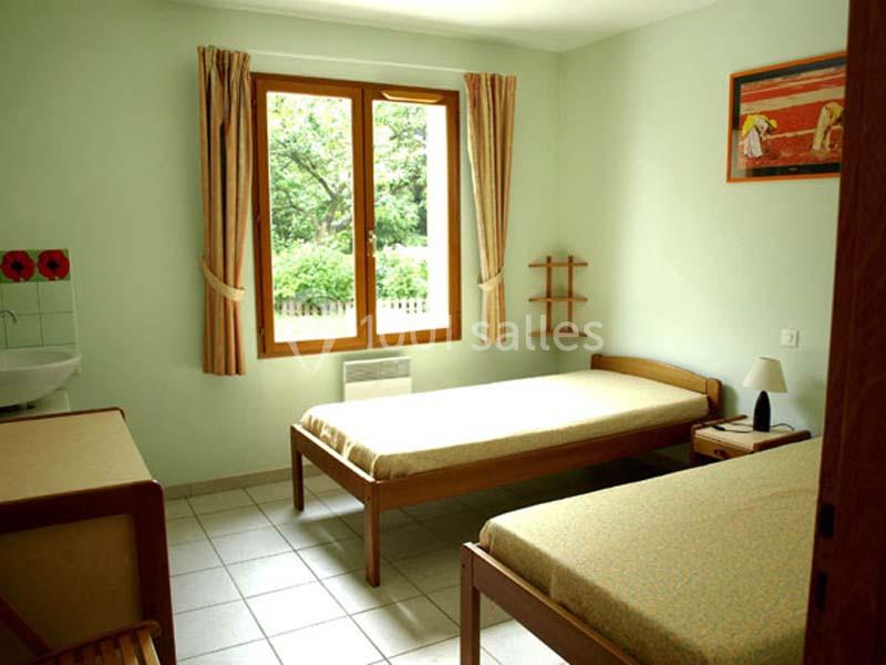 Chambre simple avec deux lits simples, une fenêtre donnant sur un jardin, des rideaux beiges et un décor minimaliste.