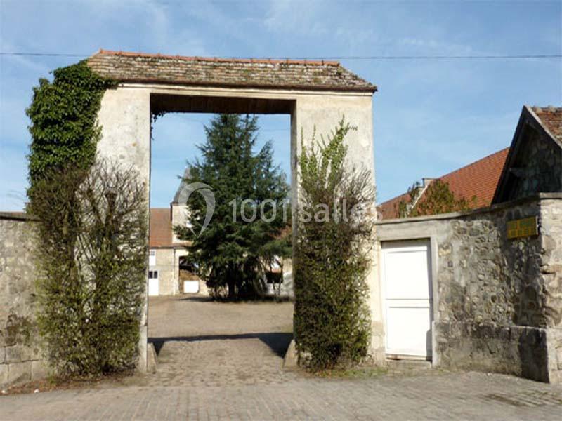 Entrée d'une cour pavée avec un portail en pierre, des murs bordés de végétation et des bâtiments en arrière-plan.