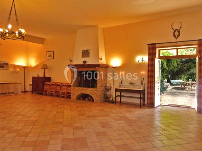 Salle spacieuse avec cheminée en pierre, mobilier en bois, carrelage au sol et accès à une terrasse extérieure verdoyante.
