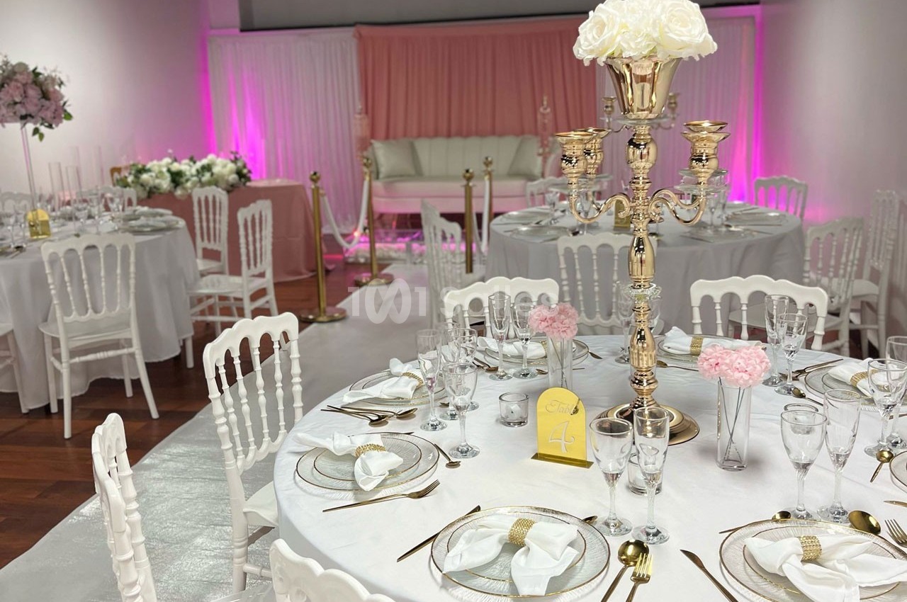 Salle de réception décorée avec des tables élégantes, des chaises blanches, un chandelier doré et un fond rose éclairé.