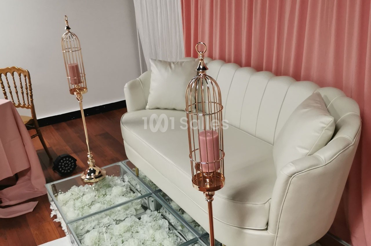 Canapé blanc devant un rideau rose, entouré de bougeoirs dorés en forme de cages et d'une table en verre.