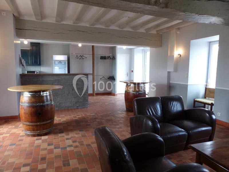 Salle avec sol en terre cuite, fauteuils en cuir, tonneaux en tables et un comptoir en pierre au fond.