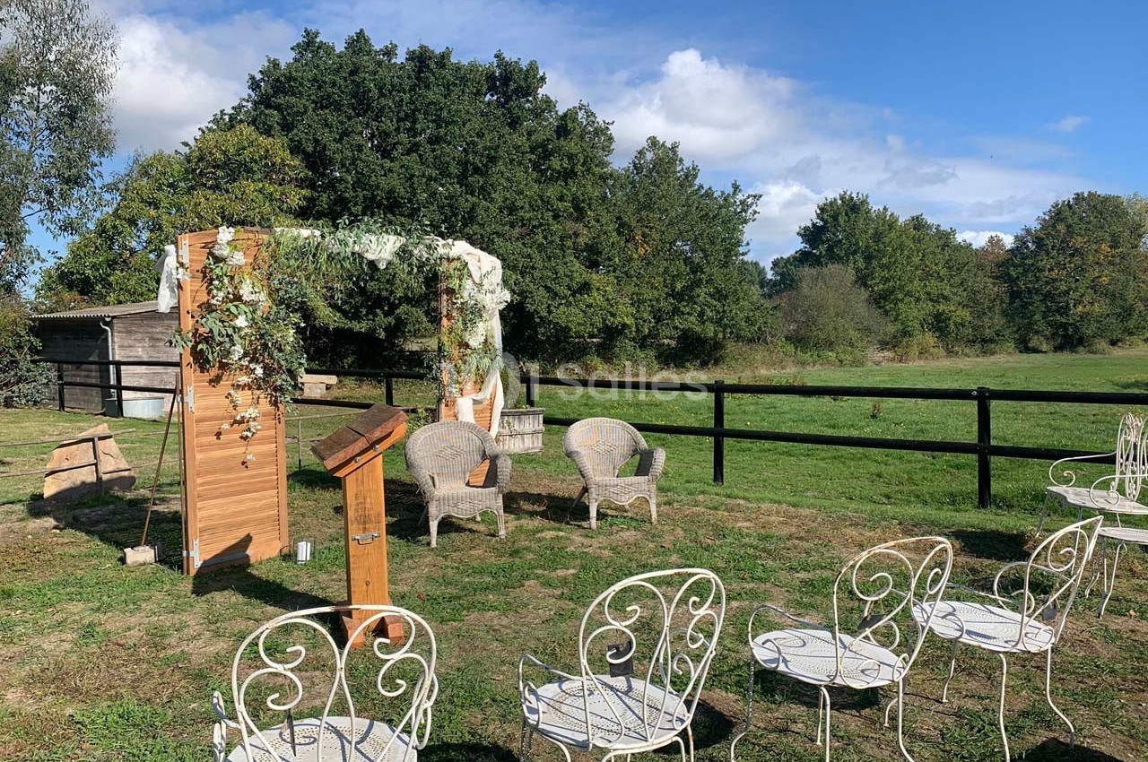 Chaises blanches disposées en extérieur devant une arche décorée de fleurs et feuillages, dans un cadre champêtre.