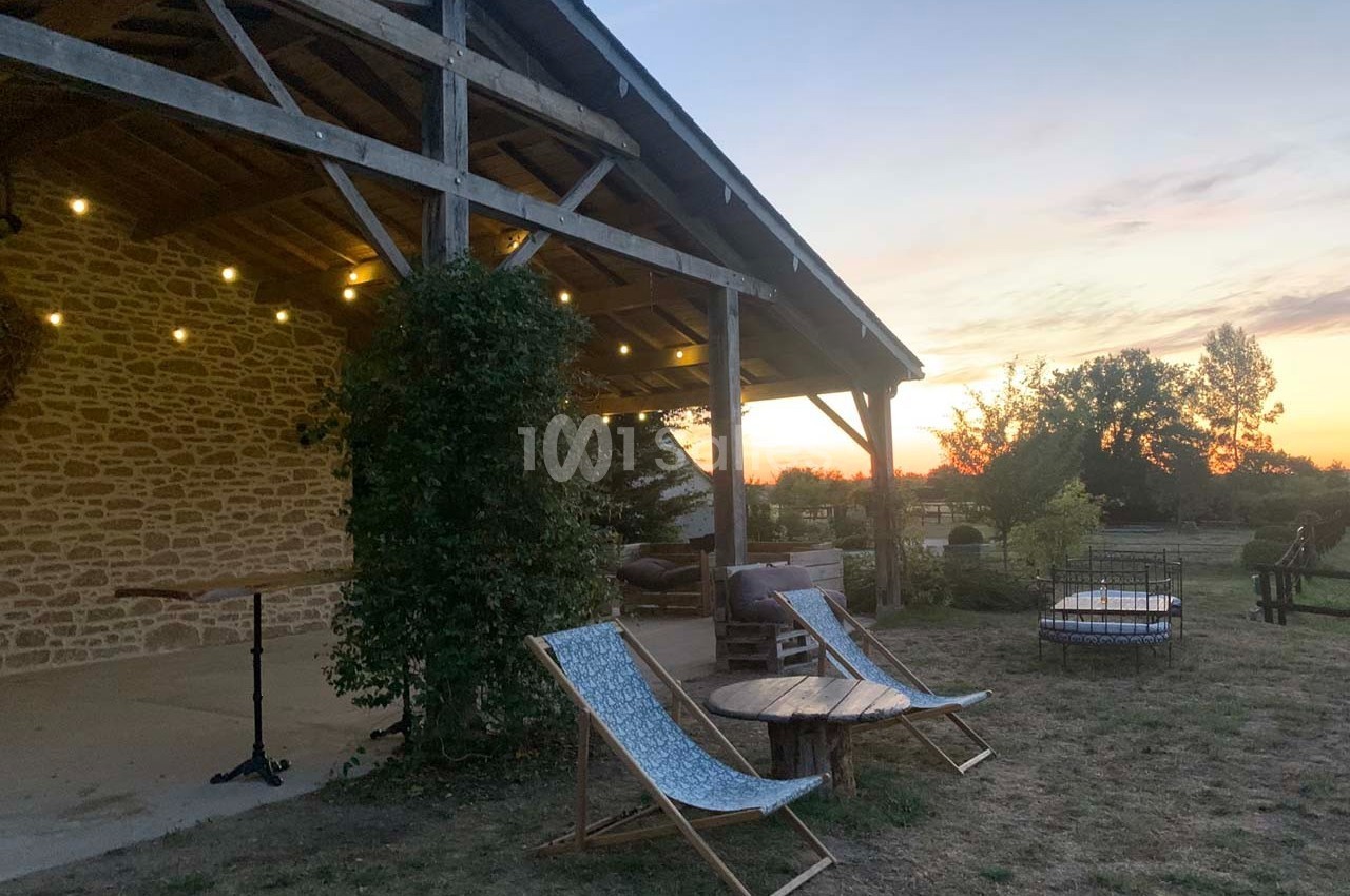 Terrasse en bois éclairée au crépuscule, avec chaises longues, table basse et vue sur un jardin arboré.