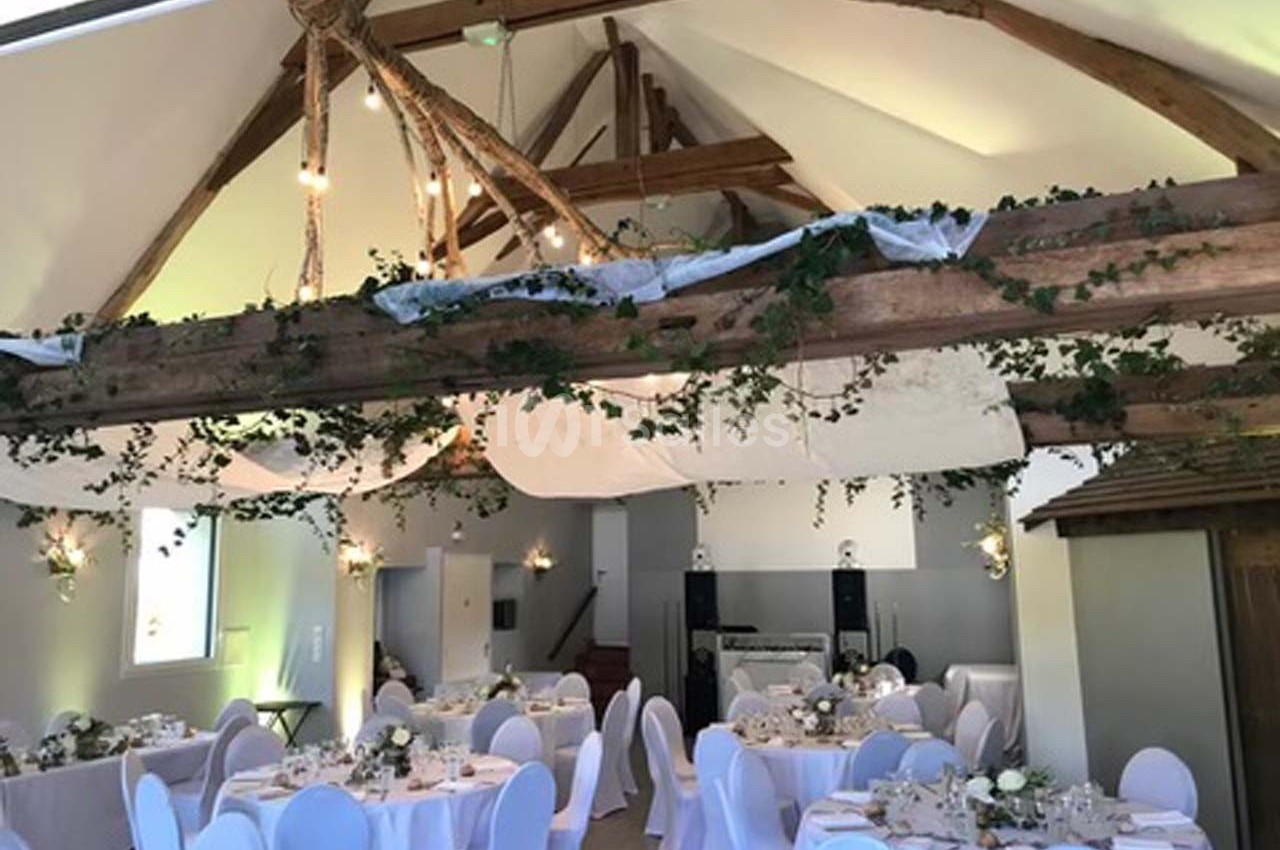 Salle de réception décorée avec des tables rondes dressées, des chaises blanches et des poutres ornées de feuillage.