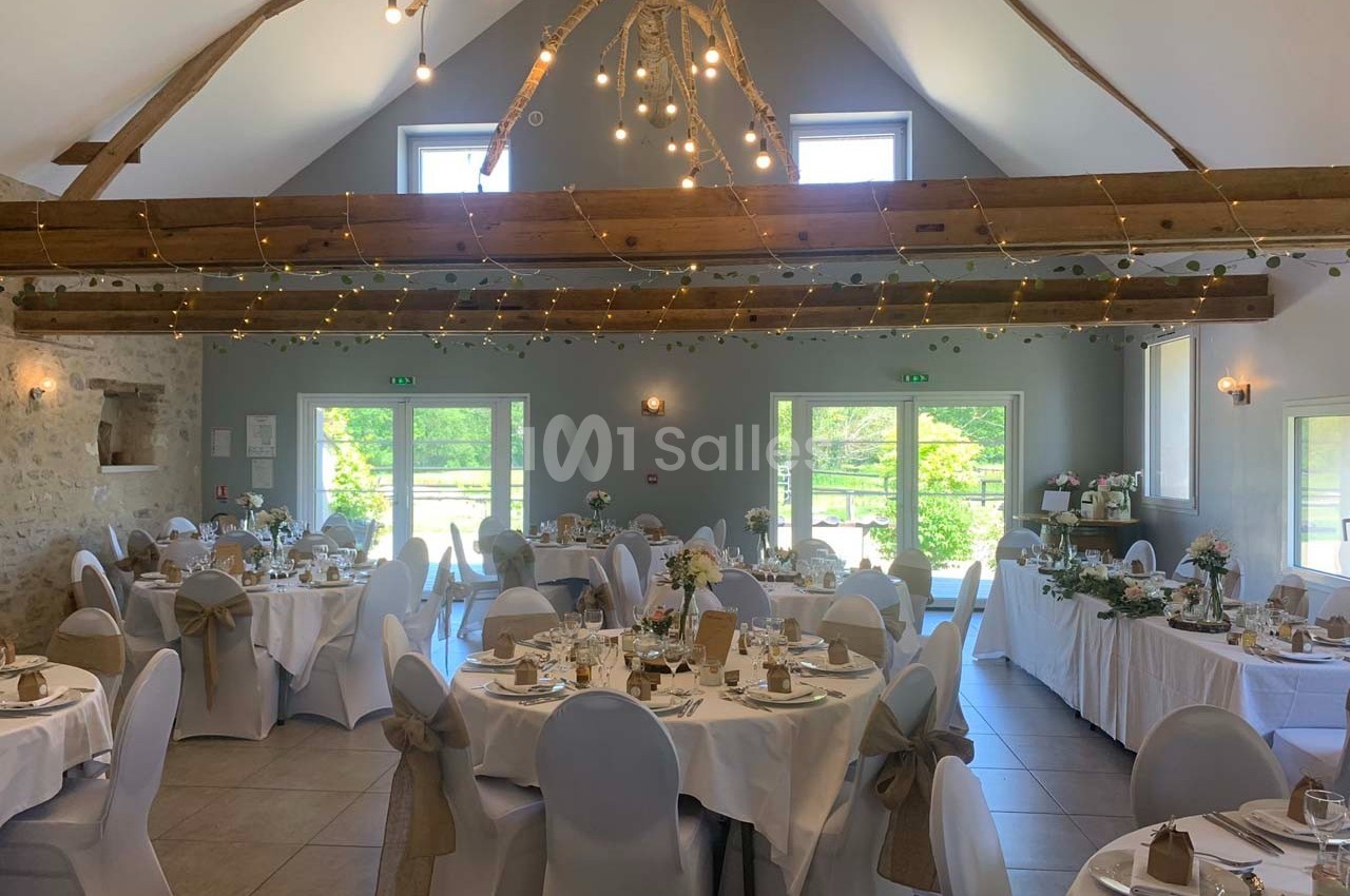 Salle de réception décorée avec des tables rondes dressées, des chaises blanches et des guirlandes lumineuses suspendues.