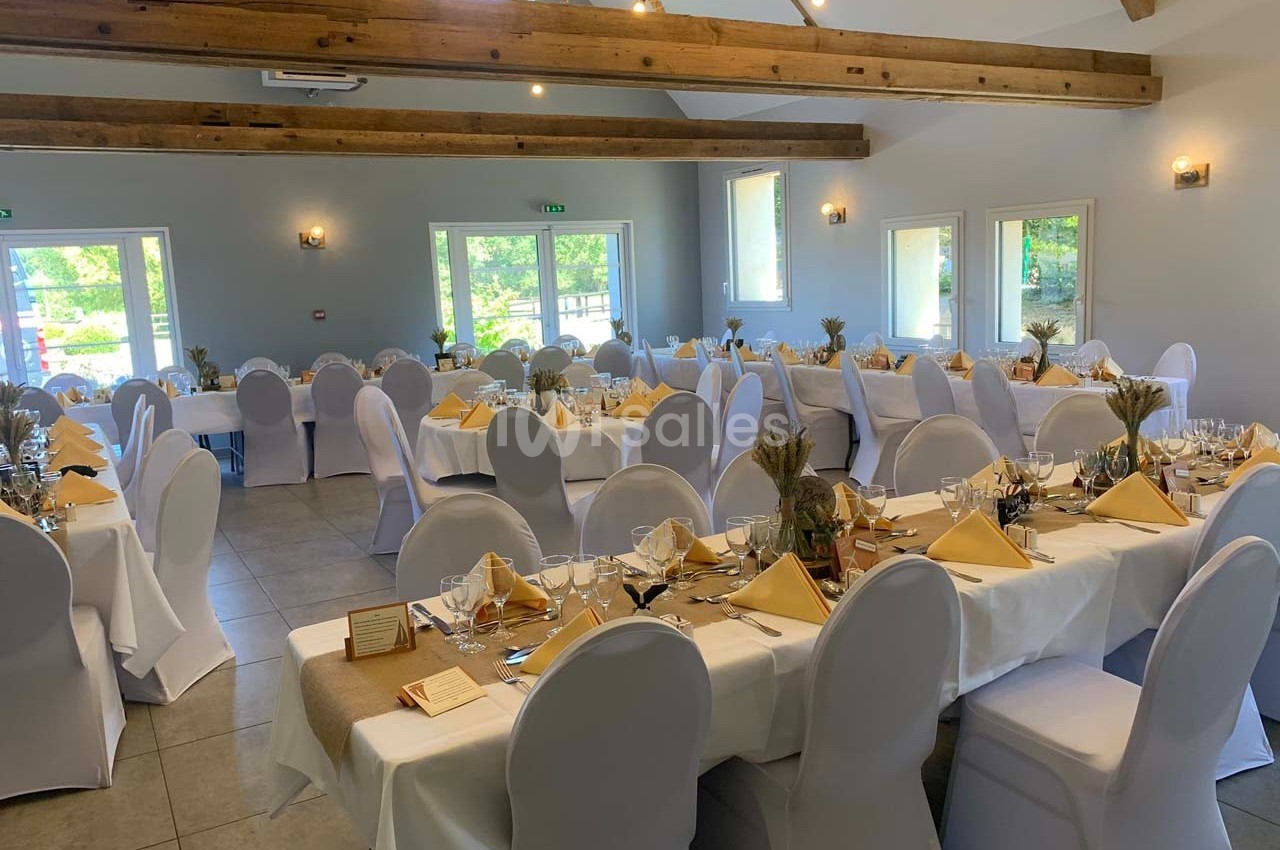Salle de réception lumineuse avec tables dressées, nappes blanches, serviettes jaunes et décoration champêtre.